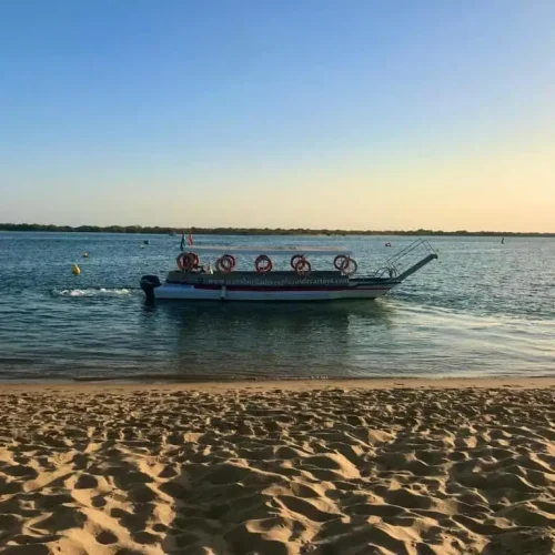 Un pequeño bote con aros salvavidas rojos en su techo flota en aguas tranquilas cerca de una orilla arenosa al atardecer, perfecto para un relajante Paseo en Barco bajo un cielo azul claro.